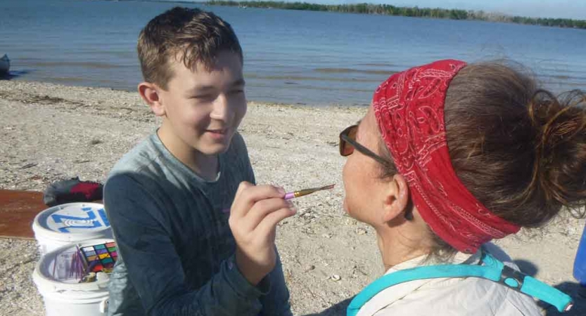 a young person paints on their parent's face on an outward bound expedition for families. They appear to be on a beach.
